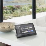 A smart hoem control tablet on the counter in a living room with a couch in the background and a view of the bay through the windows.