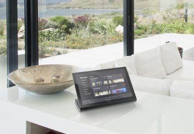 A smart hoem control tablet on the counter in a living room with a couch in the background and a view of the bay through the windows.